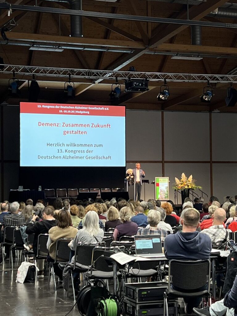 Ein Bauchredner mit Puppe steht auf der Bühne, auf der Leinwand: Demenz: Zusammen Zukunft gestalten. Herzlich willkommen zum 13. Kongress der Deutschen Alzheimer Gesellschaft.